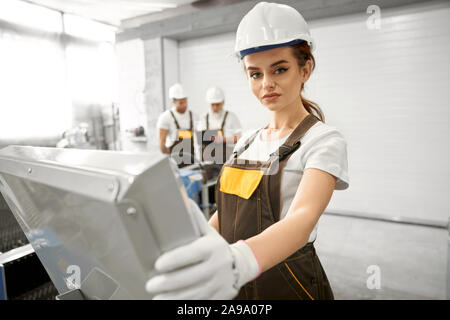 Selective focus of beautiful woman in protective mask and uniform looking at camera and posing while working with computer. Young engineer controlling equipment and devices. Concept of metalwork. Stock Photo
