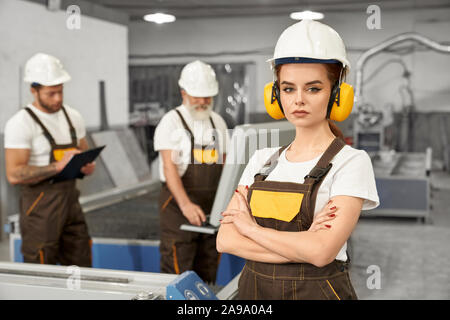 Beautiful female engineer looking at camera and posing during working day on metal factory. Young woman in helmet an uniform working with men and manufacturing products. Concept of metalwork. Stock Photo