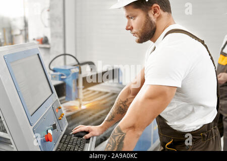 View from side of young man wearing uniform and helmet working with professional equipment and computer on factory, male welder controlling process of plasma cutting. Concept of machinery. Stock Photo