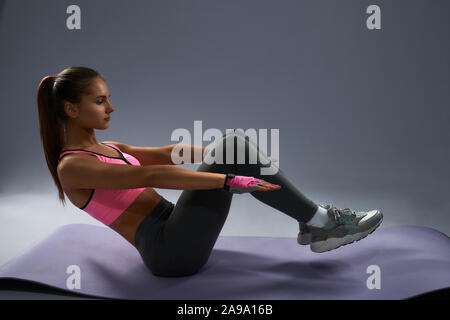 Brunette slim sporty girl with tanned skin in sportswear doing v-ups abs workout on yoga mat. Young strong female looks very concentrated while keeping balance during her daily training Stock Photo