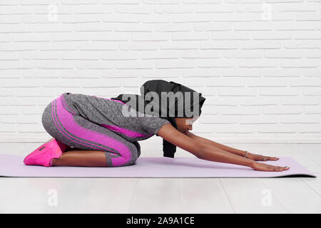 Side view of young muslim woman stretching back while doing yoga on white isolated background. Muslim religious girl in black hijab keeping fit and doing sport. Concept of health and activity. Stock Photo