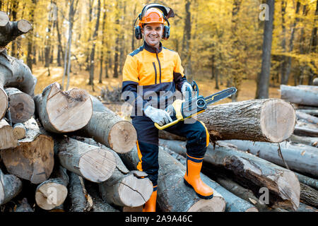 Portrait of a cheerful professional lumberjack in protective workwear standing with a chainsaw on a pile of logs in the forest Stock Photo