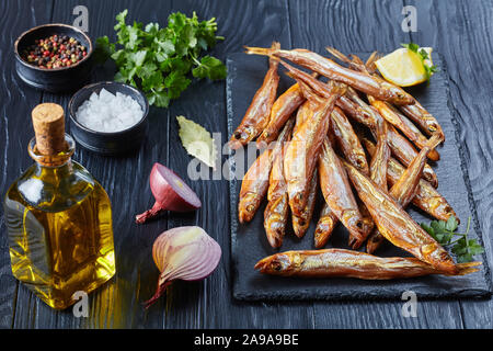 close-up of cold-smoked smelts on a black slate tray with fresh parsley and lemon wedges, on a wooden table, horizontal view from above Stock Photo