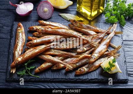 close-up of cold smoked smelts on a black slate tray with fresh parsley and lemon wedges, on a wooden table, view from above Stock Photo