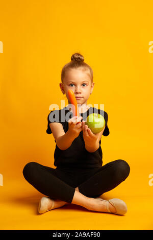 Stunned little girl in swimsuit and dance shoes, choosing healthy food and looking at camera, sitting cross-legged on a yellow background Stock Photo