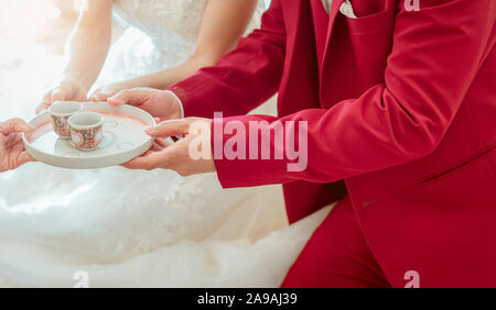 Groom and bride in tea ceremony on wedding ceremony day. Bride and groom in white wedding dress and red suit sit anf lifting tea cup. Marriage Stock Photo