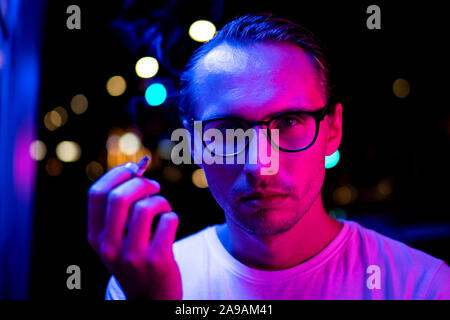 Vintage, red and blue portrait of a young man smoking a cigarette. Street lights visible in the background. Stock Photo