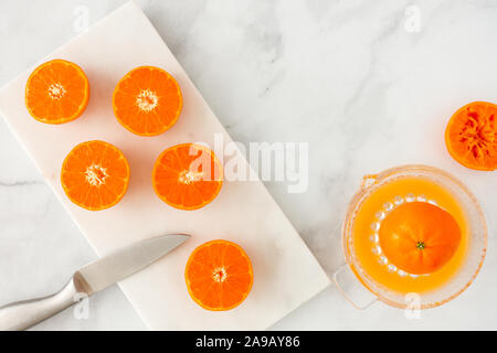 Clementines cut in halve with knife on marble chopping board and citrus juicer on white marble background with copy space. Top view. Stock Photo