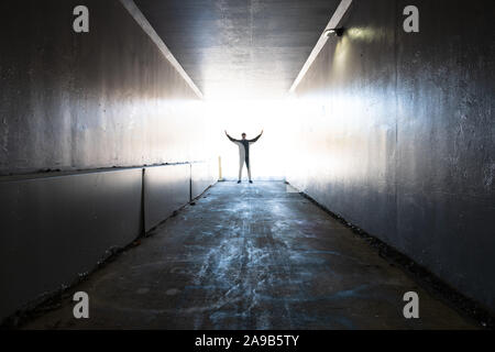 A young man stands in the entrance or end of a tunnel with backlighting in the shape of a cross. There is light at the end of the tunnel. Stock Photo
