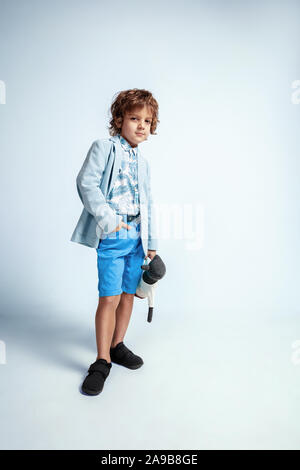 Pretty young boy in casual clothes on white studio background. Fashionable posing. Caucasian male preschooler with bright facial emotions. Childhood, having fun. Posing with his teddy bear. Stylish. Stock Photo