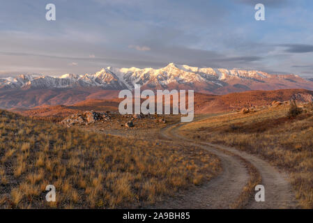 Beautiful autumn sunrise with a winding dirt road on top of a mountain against the backdrop of mountains covered with forest and snow, blue sky and cl