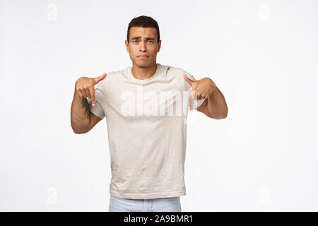 Disappointed, gloomy young tanned man in t-shirt, pointing down and looking upset, feeling displeased, distressed from seeing bad results, losing Stock Photo
