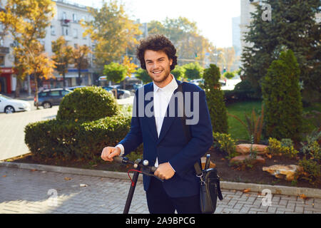 A smiling curly guy is standing with an electric scooter on a city street. Stock Photo