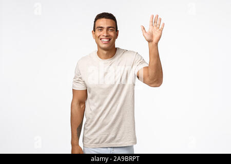 Friendly cheerful athletic man in t-shirt, raising one hand and waving in hello, hi, greeting gesture, smiling delighted and upbeat, see friend Stock Photo