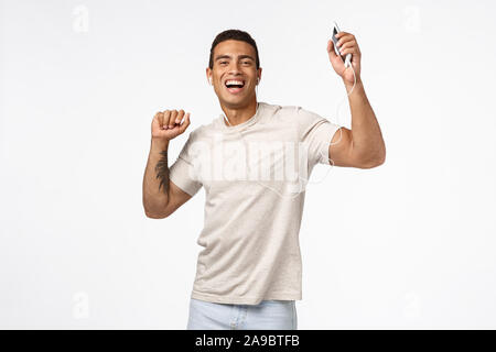 Joyful good-looking brazilian man with tattoos, raising hands up dancing carefree, enjoy awesome headphones bits, holding smartphone, wear wired Stock Photo