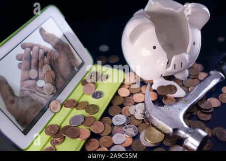 A broken piggy bank, pennies, a hammer and the image of working hands hands on the digital tablet. Stock Photo