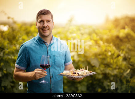 Glass of red wine in hand of a young winemaker in the vineyard - Wine tasting tour - Man welcoming tourist at sunset in the winery. Toned Stock Photo