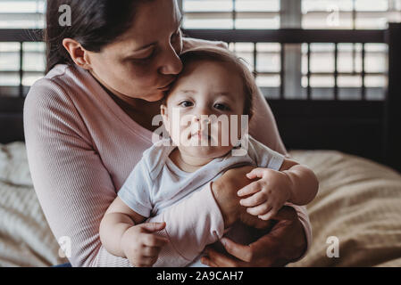 Loving mid-30â€™s mom cuddling baby girl on lap in bedroom near window Stock Photo