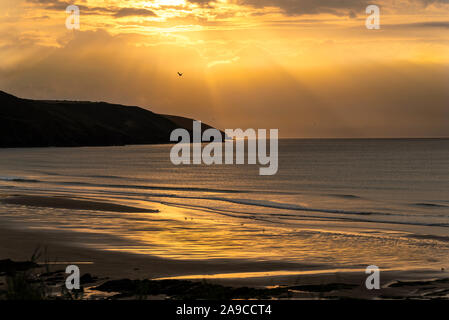 Early morning sunrise, with yellow light beams, golden light reflected on the sea surface, golden sky with clouds, headland on the horizon Stock Photo
