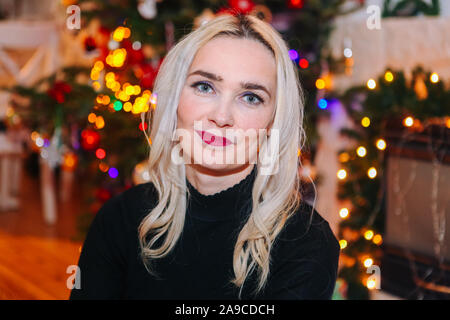 Blond woman with curly hair sit under christmas tree. New year and X-mas concept. Celebrate winter New Year holidays in decorated living room with Xma Stock Photo