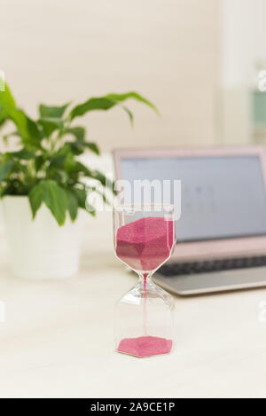 Hourglass with pink sand on a background of a laptop and a houseplant. Office time concept. Concept for time management and countdown to deadline. Stock Photo