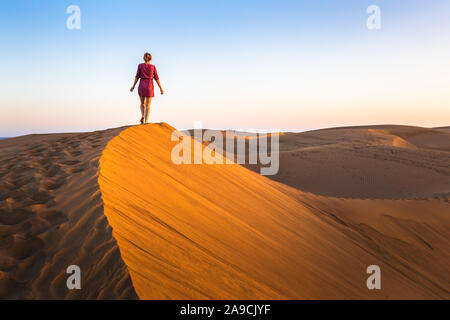 Girl walking on sand dunes in arid desert at sunset and wearing dress, scenic landscape of Sahara or Middle East Stock Photo
