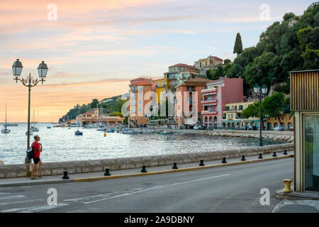 A man walks on the sidewalk next to the harbor marina at Villefranche Sur Mer, France, as the sun sets behind the colorful buildings. Stock Photo