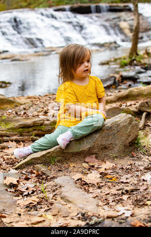 Toddler in front of waterfall sitting on slanting rock Stock Photo