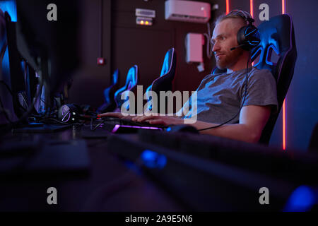 Young gamer in headphones typing on computer keyboard and looking at computer monitor while sitting on computer chair Stock Photo
