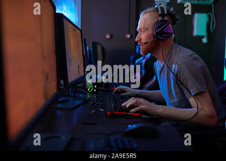 Young serious man wearing gaming headphones sitting in front of computer monitor and concentrating on his game Stock Photo