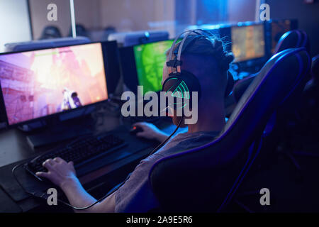 Rear view of young gamer wearing gaming headphones with backlight and playing in computer video game on computer in dark computer class Stock Photo