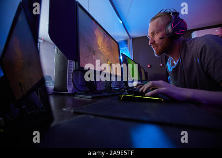 Serious young man in headphones typing on keyboard and looking at monitor attentively he playing in computer game Stock Photo