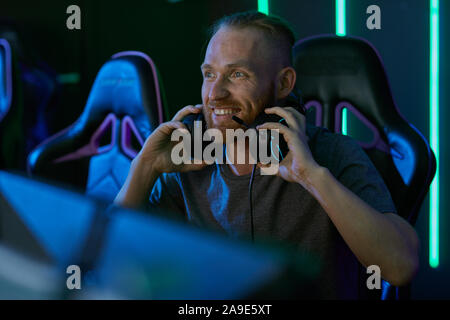 Smiling bearded man wearing headphones looking at computer monitor and smiling while sitting in dark computer class Stock Photo