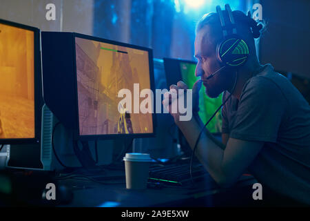 Young man in headphones sitting in front of computer monitor looking at it and thinking over the strategy of the game Stock Photo