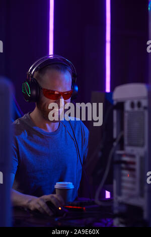 Young happy computer gamer in protective eyeglasses and in headphones sitting in front of computer monitor and playing game in dark room Stock Photo