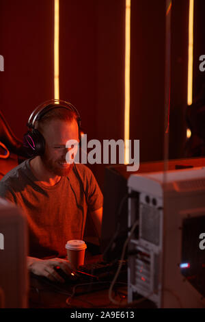 Young bearded man in headphones sitting at the table in front of computer and smiling during his work Stock Photo