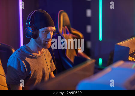 Young bearded man in hat and in headphones sitting in front of computer monitor and developing new program Stock Photo