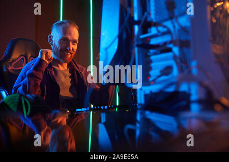 Young bearded man sitting in front of computer monitor gesturing and smiling while working in dark office Stock Photo