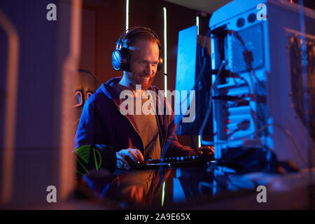 Young bearded man in headphones working on computer and smiling while sitting at his workplace in dark office Stock Photo