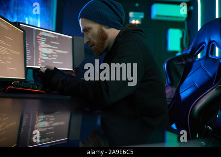 Young bearded man in dark clothing sitting at the table in front of computer monitors and trying to break into the system in dark office Stock Photo