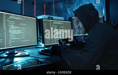 Young man in hoody using mobile phone and reading the information from computers in dark office Stock Photo