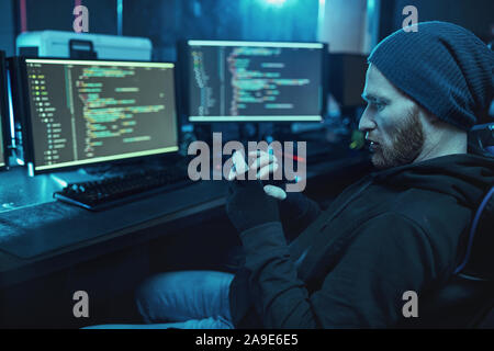 Bearded young hacker in hat and hoody shirt sitting in front of computer monitors with software and using mobile phone in dark room Stock Photo