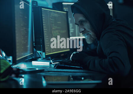 Bearded young computer hacker sitting at the table and typing on computer keyboard he breaking the system in dark office Stock Photo