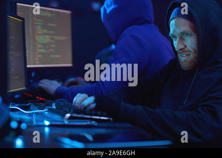 Young bearded man in hoody looking at computer monitor and typing on computer keyboard with his partner in the background in dark office Stock Photo