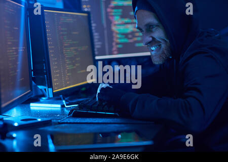 Angry computer hacker in black hood shirt enjoying the process of breaking the computer system in dark room Stock Photo