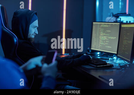 Young man in black clothing typing on computer keyboard and smiling he stealing the information from computers together with his partner Stock Photo