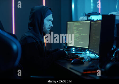 Young bearded computer hacker in hoody typing and installing the new program on computers in dark office Stock Photo