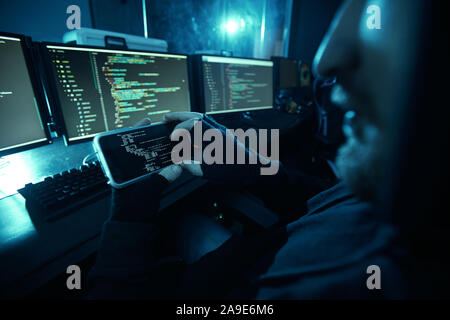 Close-up of computer hacker looking at computer monitor and guessing the password using his mobile phone in dark room Stock Photo