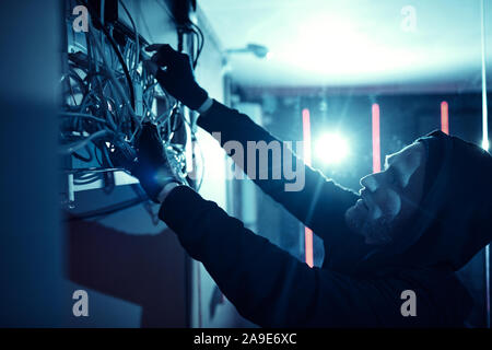 Young man in hoody standing near the box on the wall and working with cables he repairing the electricity system Stock Photo