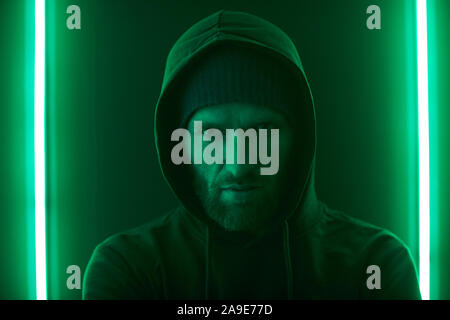 Portrait of serious bearded man in black hoody shirt looking at camera against the wall with green light Stock Photo